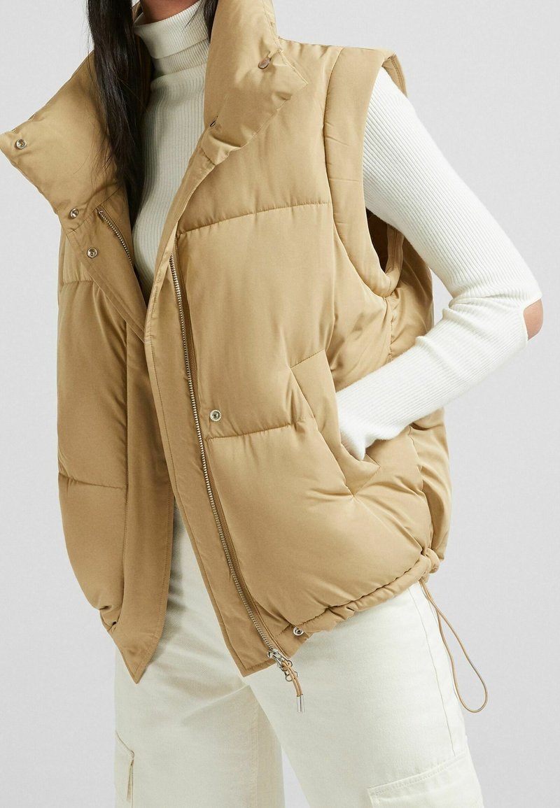 Veste matelassée beige sans manches avec un col montant, fermeture zippée et à boutons-pression, poches latérales et ourlet ajustable ; portée par-dessus un haut à manches longues côtelé blanc.