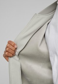 Ljusgrå zip-up jacka delvis öppen, vilket avslöjar en slät, mjuk innerfoder. En hand håller i kanten, vilket visar detaljer i tygstrukturen.