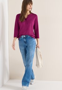 Femme marchant à l'intérieur portant une chemise à manches longues magenta, un jean bleu, des baskets argentées, tenant un pull beige dans une main.