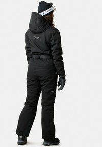 Zwarte skisuit met capuchon en verstelbare riem, met een gestructureerde afwerking en logo-detail op de rug, gecombineerd met zwarte handschoenen en laarzen.