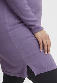Tunic a maniche lunghe viola con una texture liscia, dotata di spacchi laterali all'orlo e maniche aderenti, abbinata a leggings neri.