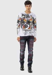 Langärmliges weißes Hemd mit grafischen Drucken in Schwarz und Rot; abgewetzte dunkle Jeans mit roten Akzenten und Flicken; abgetragene schwarze Stiefel.