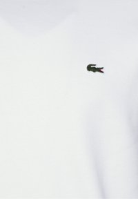 Valkoinen kangas, jossa on pieni brodeerattu vihreä krokotiililogo punaisella suulla vasemmalla ylhäällä.