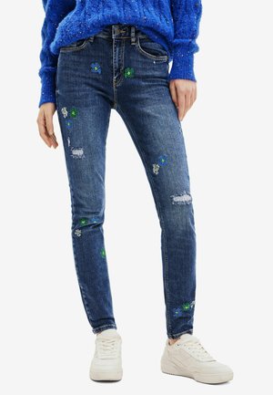 Niebieskie jeansy w fasonie slim fit z małym kwiatowym haftem i przetarciami, noszone z białymi trampkami i niebieskim swetrem.