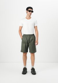 Mann med svarte solbriller, hvit t-skjorte, grønne shorts, grå sokker og svarte joggesko som står med hendene i lommene mot hvit bakgrunn.