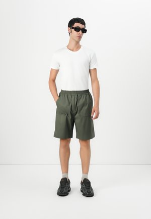 Man in zwarte zonnebril, witte t-shirt, groene shorts, grijze sokken en zwarte sneakers, met zijn handen in de zakken tegen een witte achtergrond.