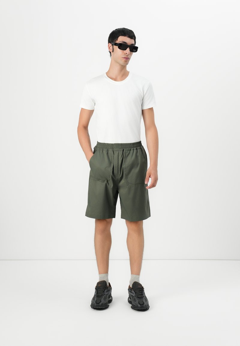 Bărbat îmbrăcat cu ochelari de soare negri, tricou alb, pantaloni scurți verzi, șosete gri și adidași negri, stând cu mâinile în buzunare pe un fundal alb.