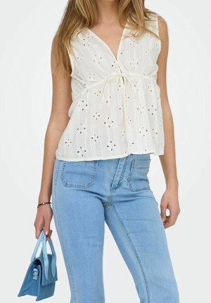 Witte top met korte mouwen, voorzien van uitgesneden bloemenborduursels en een taillering, gecombineerd met lichtblauwe jeans en een kleine blauwe handtas.