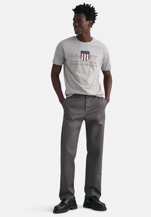 Grå bomulds t-shirt med et skjoldlogo i marineblå og rød, parret med løstsiddende grå bukser og sorte snørebåndssko.