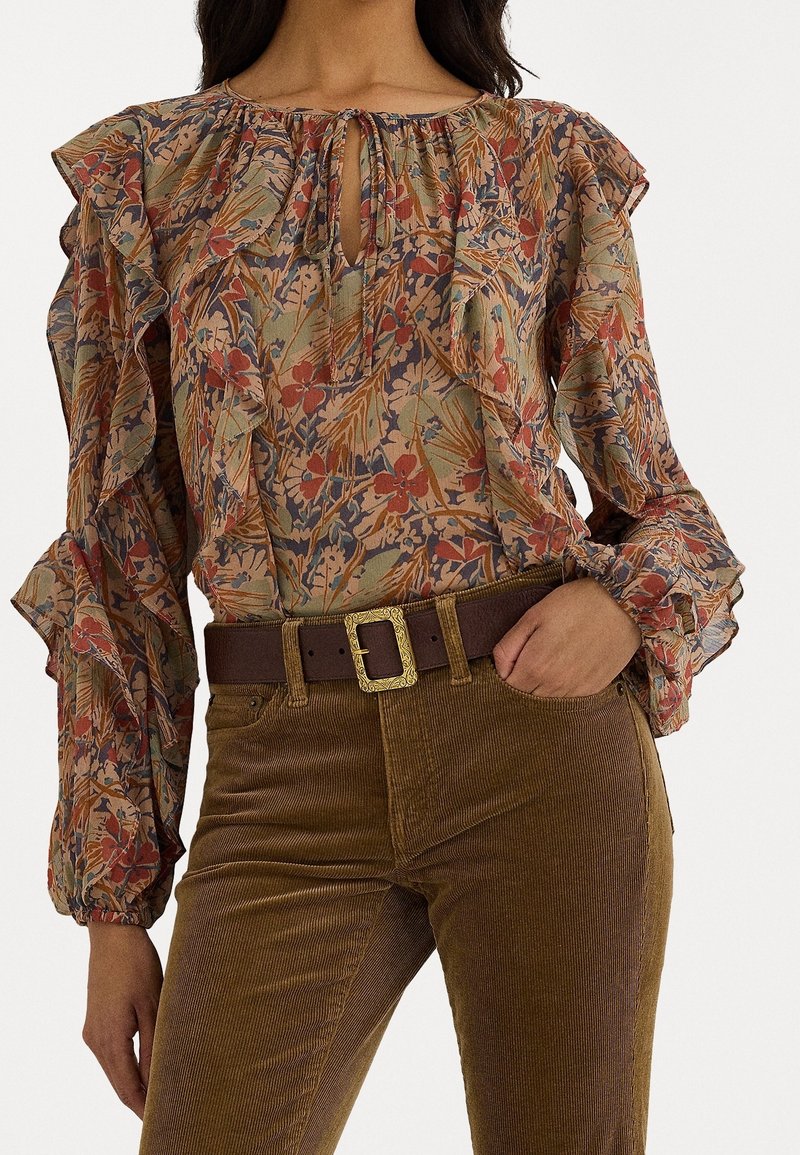 Femme portant un chemisier fleuri à manches longues avec des volants et un pantalon en velours marron avec une large ceinture marron à boucle dorée.