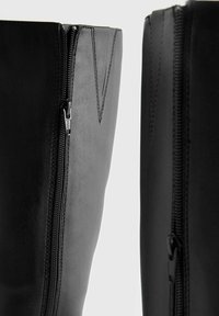 Bottes noires en cuir haute jusqu'au genou avec une texture lisse, dotées d'une fermeture éclair latérale et de détails en surpiqûre pour plus de caractère.