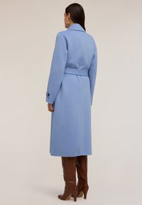 Jasnoniebieski wełniany płaszcz z paskiem w talii, wyciętym kołnierzem i guzikami na mankietach. Gładka tekstura i długość do kolan, noszony na brązowe botki na wysokim obcasie.