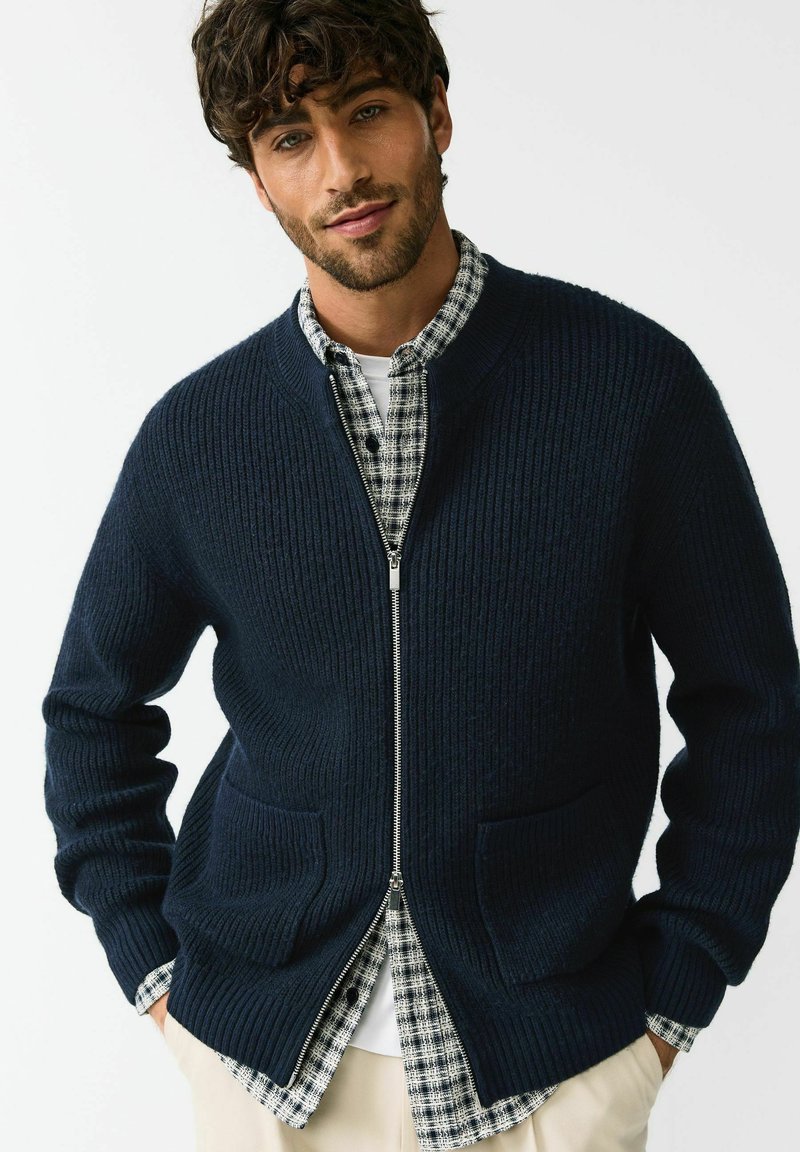 Cardigan bleumarin din tricot cu dungi, prevăzut cu fermoar și două buzunare frontale. Purta peste o cămașă în carouri, evidențiind un aspect stratificat.