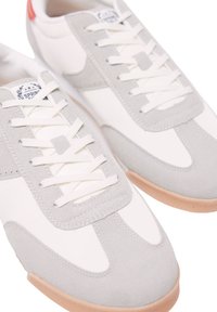 Weiße und graue Sneaker mit Obermaterial aus Wildleder und Stoff, Schnürdesign, Gummisohle und Logopatch auf der Zunge. Roter Akzent an der Ferse.