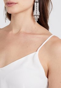 Femme portant un haut sans manches blanc et de longues boucles d'oreilles en argent en forme de lustre avec des détails géométriques et étoilés.