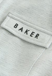 Harmaa kangas, jossa on hieno ruutukuvio; siinä on etiketti, jossa lukee "BAKER" paksuilla mustilla kirjaimilla. Pintakuvio näyttää sileältä ja kudotulta.