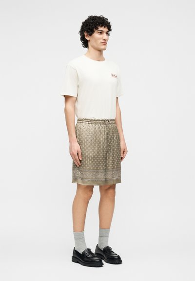 Jeune homme aux cheveux bouclés portant un t-shirt blanc, un short à motifs arrivant aux genoux, des chaussettes grises et des mocassins noirs, debout devant un fond blanc.