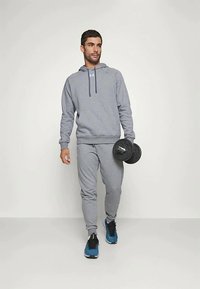 Bărbat îmbrăcat într-un trening gri și adidași albaștri, mergând în timp ce ține un ganteră neagră în mână dreaptă, pe un fundal alb simplu.