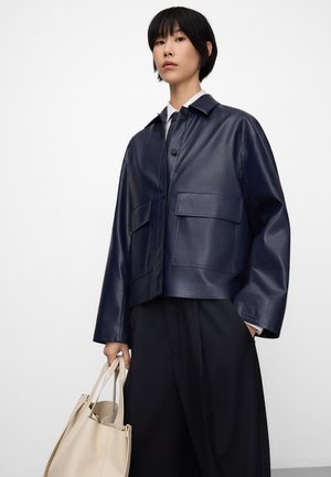 Osoba ubrana w ciemnoniebieską skórzaną kurtkę na białej koszuli i czarnych spodniach, trzymająca beżową torbę na ramię jedną ręką.