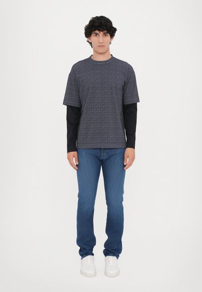 Kratkās piedurknes iešūts tumši zils T-krekls virs melna garām piedurknēm krekla, saskaņots ar šauriem ziliem džinsiem un baltiem sporta apaviem.