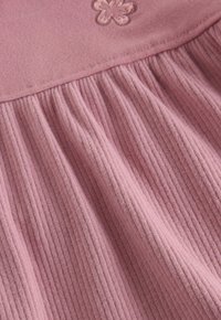 Rosa Stoff mit gerippter Textur, der ein gerafftes Design und ein gesticktes Blütenmotiv nahe dem oberen Rand aufweist. Weiches und leichtes Material.