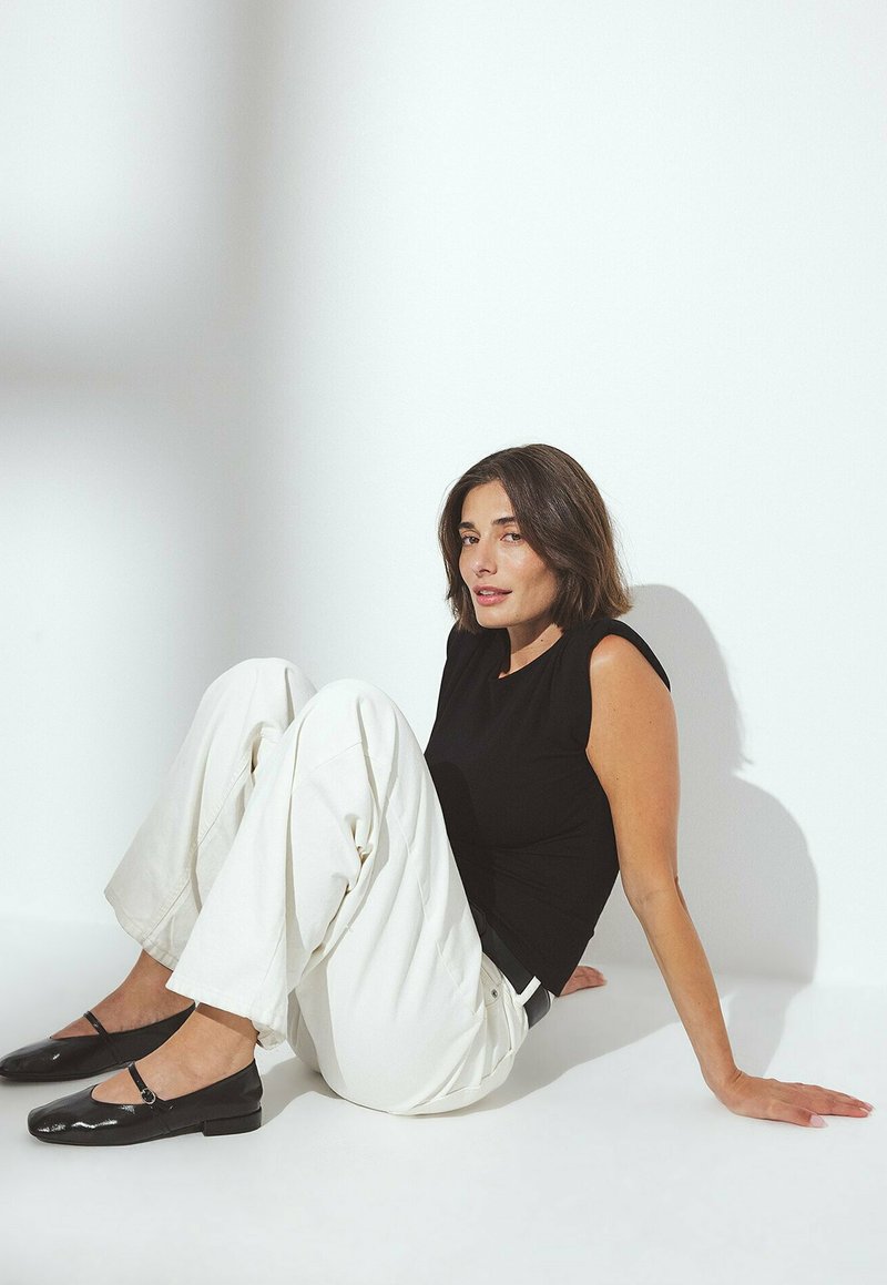 Top nero senza maniche, pantaloni bianchi larghi e ballet flats neri in pelle lucida. Sfondo neutro, posa seduta, texture lisce, accentuazioni minime.