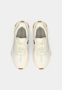 Ζευγάρι αθλητικών παπουτσιών σε κρεμ και μπεζ χρώμα με ελαστικά κορδόνια τύπου μπαλόνι, γκρι εσωτερικές σόλες και καφέ λεπτομέρειες, φαίνεται από πάνω σε λευκό φόντο.