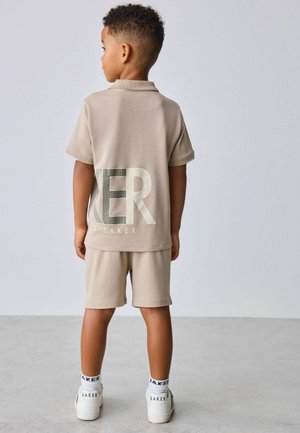 Beežist lühikeste varrukatega polo-särk, millel on suured helkurkirjad seljal. Kandes sobivate beežide lühikeste pükstega ja valgete tossudega.