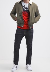 Olivgrüne Bomberjacke mit beigem Lammfellkragen, roter Pullover mit weißem Rentiermuster, dunkle Jeans und wei�ße Sneakers mit schwarzen Streifen.
