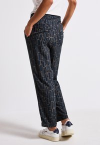 Modella che indossa pantaloni navy a fantasia con una vestibilità comoda e tasche, abbinati a sneakers bianche, in piedi contro uno sfondo semplice.