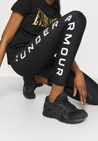 Osoba w czarnych legginsach Under Armour i czarnych butach do biegania Asics, klęczy z jedną ręką blisko ziemi.