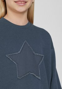 Šedá mikina s velkou, třep�ící se hvězdou vyšitým na přední straně. Měkká textura a kulatý výstřih, s klesajícími rameny.