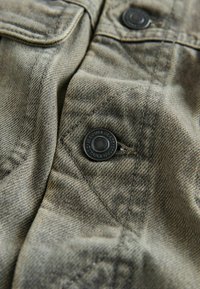 Izbalējis olīvu zaļš džinsa materiāls ar teksturētu virsmu, aprīkots ar melnu spiedpogu un izšūtu kabatas detaļu.