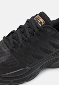 Närbild av en svart sportskor med texturerat nät och släta sektioner, med synliga snören och en logotagg på tungan.