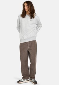 Sudadera gris de cuello redondo con un pequeño logo negro, combinada con pantalones marrones holgados y zapatillas en blanco y negro con acentos texturizados.