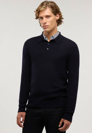 Joven con cabello rubio ondulado que lleva un suéter texturizado azul marino oscuro sobre una camisa a rayas en blanco y negro con cuello.