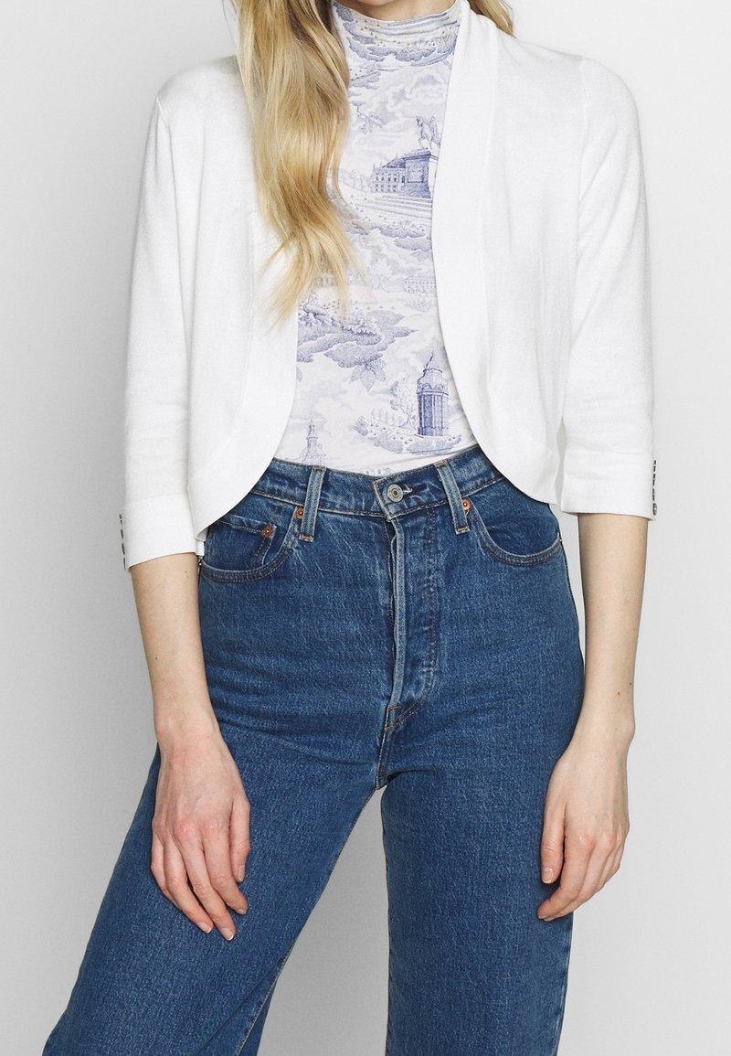 Cardigan bianco, leggero, con maniche a tre quarti, abbinato a un top leggero a fantasia lilla e jeans blu a vita alta.