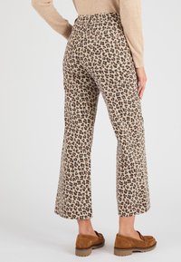 Pantalones acampanados beige con un diseño de estampado de leopardo en negro y marrón, combinados con zapatos de ante marrones tipo slip-on. La tela parece suave con un acabado texturizado.