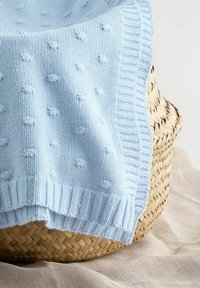 Svetlomodrá pletená deka s vystupujúcim vzorom bodiek, prehodená cez tkaný prútený kôš na béžovom látkovom povrchu.