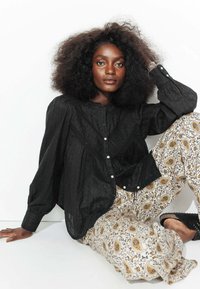 Blouse noire à texture avec fermeture à boutons, associée à un pantalon crémeux large avec un motif floral paisley jaune et noir.