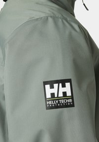 Chaqueta verde resistente al agua con una textura suave y un distintivo parche de logo negro marcado "HELLY TECH® PROTECTION."