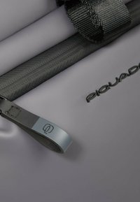 Bolsa impermeable gris en primer plano con cremallera negra y asa, con logo en relieve y textura suave.