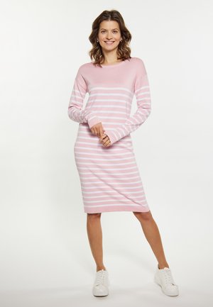 Kvinde smilende iført en pink og hvid stribet langærmet kjole og hvide sneakers, stående mod en ensfarvet lys baggrund.