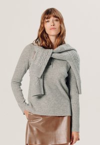 Grijze geribbelde trui met een kabelgebreide sjaal om de nek. Bruine neplederen rok. Zachte texturen en een getailleerd ontwerp.