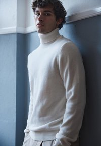 Young man with dark curly hair wearing a white turtleneck sweater stands against a blue and white wall indoors.