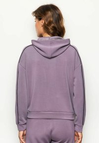 Mujer con cabello castaño ondulado, vistiendo una sudadera color lavanda y pantalones a juego, mirando hacia atrás contra un fondo claro y liso.