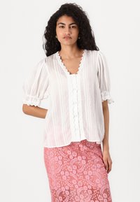 Blouse blanche légère avec un décolleté en V festonné, des manches bouffantes et une fermeture par boutons. Tissu texturé avec des rayures verticales.
