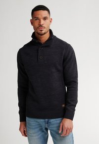 Man in een zwarte gebreide pullover trui met een knoopsluiting en lichtblauwe jeans voor een effen achtergrond.