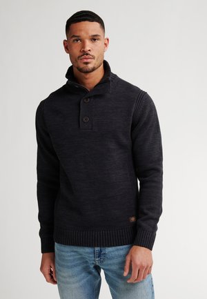Hombre vistiendo un suéter de punto negro con un cuello abotonado y jeans azul claro frente a un fondo liso.