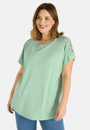 Lächelnde Frau in einem lockeren mintgrünen Oberteil mit floralen Spitzen-Aussparungen an den Schultern und blauer Jeans vor einem weißen Hintergrund.